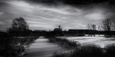 Ice and snow covered river, trees, rushes, fields and cloudy sky. Cold, snowy winter landscape. Monochrome black and white. Shades of winter
