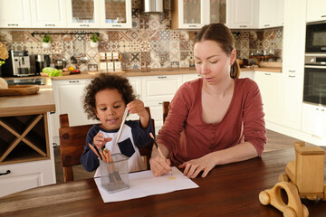 Cute little biracial boy taking crayon while his mother drawing picture on paper by wooden kitchen table - Powered by Adobe