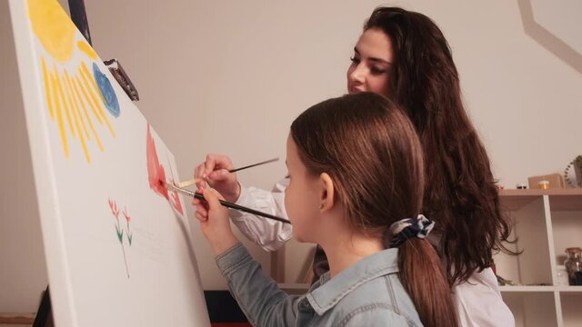 Art studio. Kids creativity. Painting lesson. Little girl drawing scenery picture together with female teacher on white canvas in light room interior.