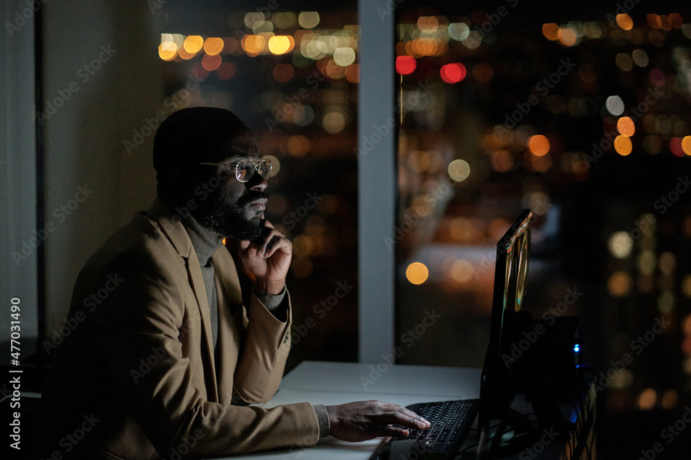 Poster young serious african businessman looking through online data while sitting in front of computer mon