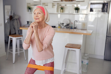 Active aged lady working out while doing squats with elastic band on blurred interior of modern flat