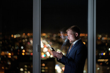 Side view of businessman in formalwear scrolling in mobile phone against large office window while working at night