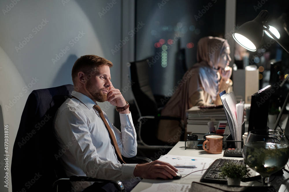 Poster young bearded businessman concentrating on work while sitting by desk in front of computer late at n