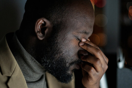Face of exhausted African businessman touching his closed eyes while sitting by workplace in office at night