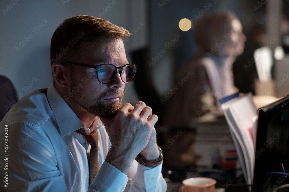 Poster serious businessman in formalwear and eyeglasses sitting in front of computer screen late in the nig