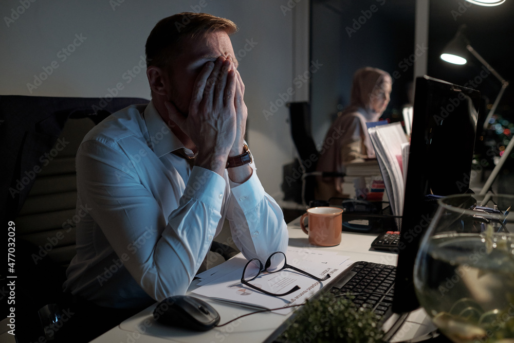 Poster exhausted businessman in white shirt keeping his arms on face while working in front of computer in 