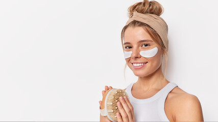 Horizontal shot of beautiful smiling European woman applies beauty patches under eyes wears headband and t shirt holds body brush poses against white background copy space for your promotion