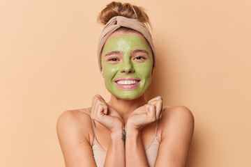 Happy young European woman keeps hands under chin smiles gladfully applies green nourishing mask...