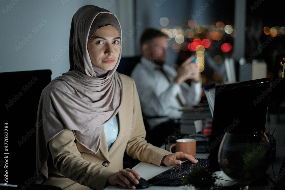Poster contemporary young businesswoman in hijab looking at camera while sitting in front of computer in of