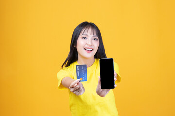 Portrait Asian happy young girl smiling cheerful and showing plastic credit card while holding mobile phone isolated on yellow background