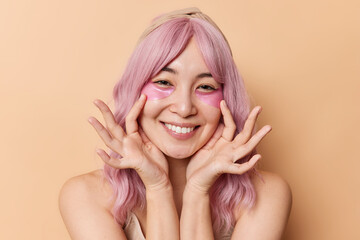 Happy beautiful pink haired young Asian woman applies hydrogel patches under eyes to reduce wrinkles smiles happily enjoys skin care treatments poses with bare shoulders against beige background.