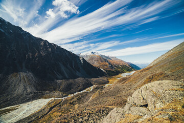 Picturesque highland landscape with mountain river and lake in valley among high mountains with snow in autumn colors in sunshine. Awesome alpine view to motley mountain valley under cirrus clouds.