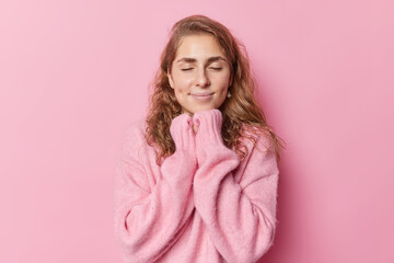 Peaceful calm beautiful woman with wavy hair pleasant appearance keeps hands under chin enjoys cozy atmosphere at home during cold winter day wears warm soft jumper poses indoor against pink wall