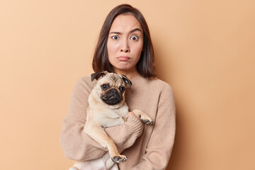 Shocked surprised brunette woman raises eyebrows has stunned expression poses with pug dog spends free time with favorite pet wears casual jumper isolated over beige background. Animals concept