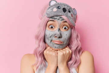 Surprised Asian woman keeps hands under chin applies nourishing mask on face has wondered expression undergoes beauty treatments before sleep has long pink dyed hair wears sleepmask poses indoor