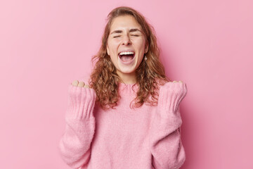 Euphoric overjoyed long haired woman keeps mouth widely opened exclaims from joy clenches fists with triumph celebrates achhievement wears warm sweater isolated over pink background reaches goal - obrazy, fototapety, plakaty