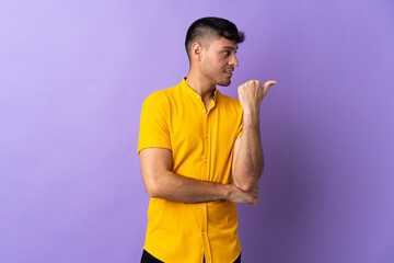 Young Colombian man isolated on purple background pointing to the side to present a product