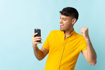 Young Colombian man using mobile phone isolated om blue background celebrating a victory