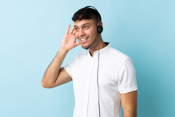 Telemarketer Colombian man working with a headset over isolated background listening to something by putting hand on the ear