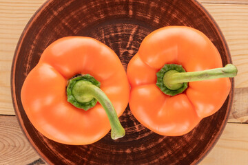 Two orange fresh sweet peppers with a clay dish on a wooden table, close-up, top view.