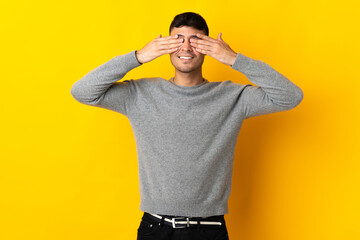 Young Colombian man isolated on yellow background covering eyes by hands and smiling