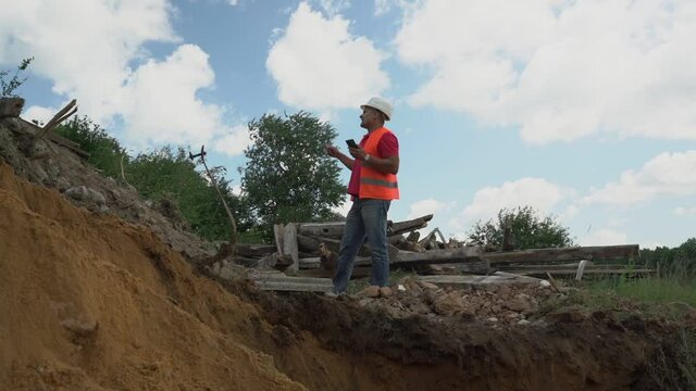 adult male foreman in hard hat and retroreflective vest stands on edge of trench on construction site and gives instructions for excavator driver