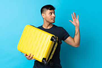Young Colombian man isolated on blue background in vacation with travel suitcase and saluting