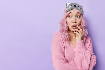Photo of surprised pink haired young woman looks embarrassed aside reacts on something awesome wears blindfold and jumper hears shocking news isolated over purple background copy space area.