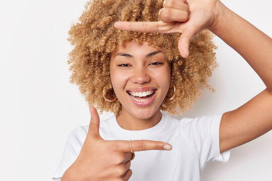 Snap. Positive woman with curly hair takes picture looks through hand frame as if photographing with camera smiles gladfully isolated over white background captures moment pictures something in mind