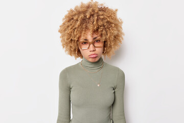 Upset woman with curly hair purses lips and looks sadly at camera has problems wears spectacles and...