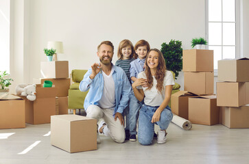 Fototapeta na wymiar Happy family showing keys to their new home. Portrait of mom, dad and little children looking at camera and smiling in spacious living room full of cardboard boxes. Buying house or apartment concept