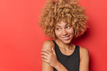 Curly haired young woman keeps hand on shoulder dresse in black t shirt concentrated away with dreamy expression isolated over red background with copy space. Pleasant face expressions concept