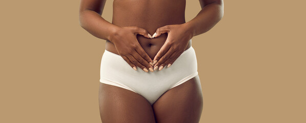 Plus size black woman in underwear holding hands on abdominal area forming heart shape around navel, isolated on solid brown color background, cropped shot. Self love, care, health, pregnancy concept © Studio Romantic