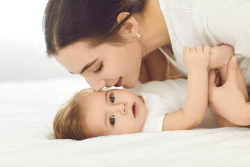Mother and child connection. Portrait of happy young mother tenderly kissing baby girl lying on white bed at home. Close up side view of mom with baby playing and spending time together.