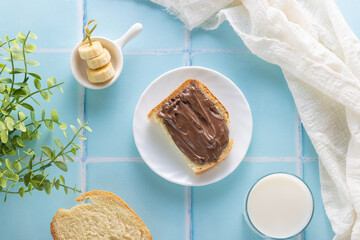 a sandwich with chocolate paste, milk and banana slices on a background of blue tiles and a green branch. Delicious breakfast, flat lay, top view