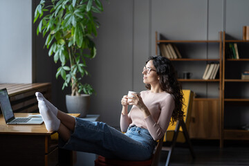 Young woman relaxing at office or cozy home workplace with bare feet on desk drinking tea or coffee. Happy satisfied millennial female freelancer rest sitting at laptop think of future success concept