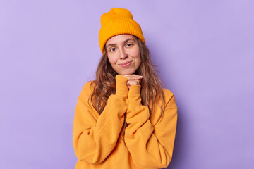 Charming long haired woman looks with tender expression keeps hands under chin wears casual orange jumper and hat isolated over purple background. People face expressions and felings concept