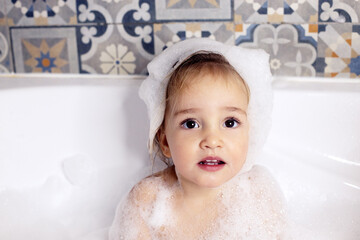 Cute kid with big beautiful eyes sitting in bathroom with bath foam. Baby care, personal care. Girl