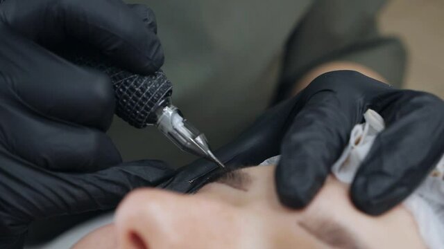 The master makes permanent eyebrow makeup with a needle tattoo machine.The procedure for applying the pigment using special equipment.The procedure is carried out in gloves. Eyebrow microblading