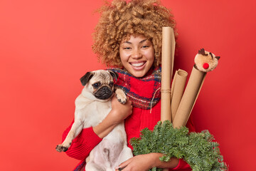 Happy woman holds pug dog celebrates festive event carries holiday attributes decorates room before...