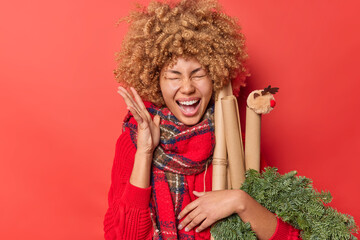 Indoor shot of overjoyed emotional curly haired woman exclaims loudly keeps hand raised has happy expression anticipates winter holidays poses against red background. Yeah New Year is coming