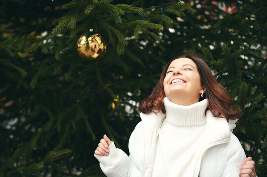 Outdoor portrait of happy woman next to green Christmas tree, wearing white pullover and jacket