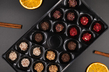 Colorful chocolate truffles in a candy box with cinnamon and dried oranges on a black background. Top view.