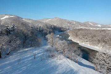 A frozen river flows between trees covered with white snow. On a frosty sunny December day in a...