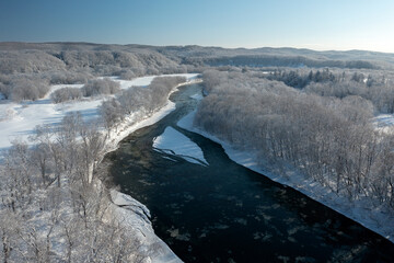 A frozen river flows between trees covered with white snow. On a frosty sunny December day in a...