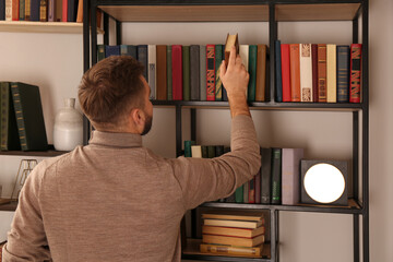 Young man choosing book on shelf in home library, back view