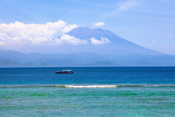 Mount Agung active volcano covered by clouds in Nusa Penida, Bali island, Indonesia. Traditional fishing boats called jukung on the white sand beach. High waves with foam spread on the coast.