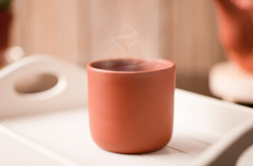 Earthenware mug with hot steam, coffee or tea on a white wooden tray, with partial view of the kettle, brown wooden background.