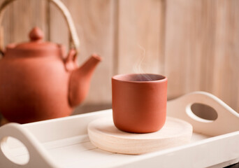 Earthenware mug with hot steam, coffee or tea on a white wooden tray, with partial view of the kettle, brown wooden background.