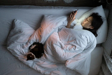 Lazy unmotivated woman staring at smartphone screen while lying in bed with cat at night, female being addicted to social media, feeling depressed, suffering from sleep desorder. Life apathy concept